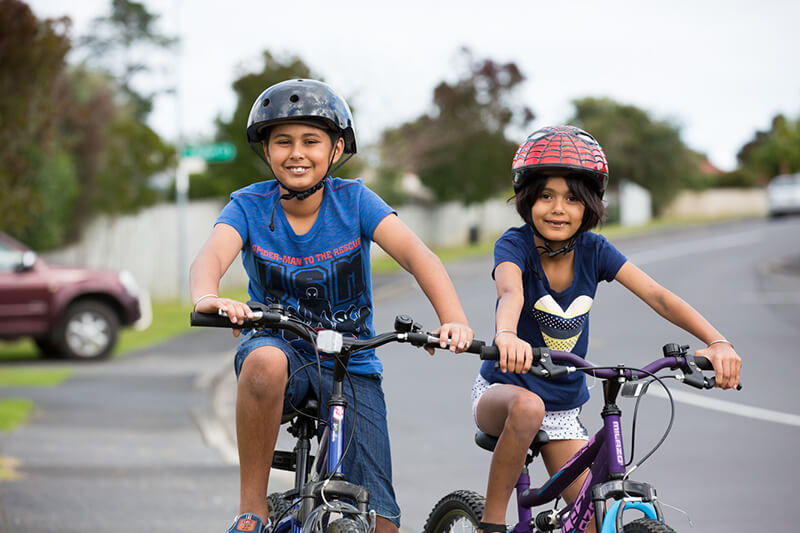 two children on their bikes outdoors. Both are wearing bike helmets and smiling at the camera.