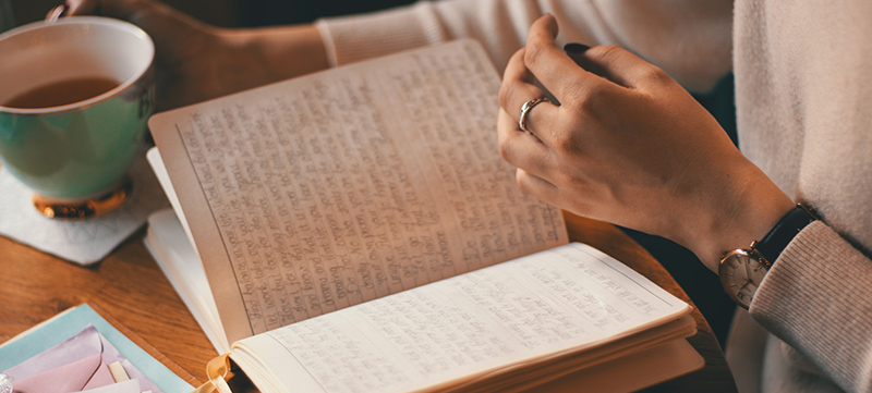 Close up of a book open to pages of hand-written text. A cup of coffee is held in the background